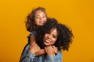 A mother with styled hair and a jean jacket smiles and holds her smiling daughter on her shoulders while posing in front of an orange background