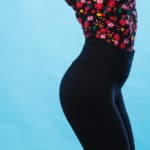 A close up of a woman's body wearing black pants and a floral tucked in blouse in front of a blue background